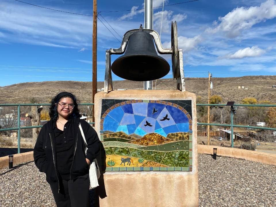 Aspen Coriz-Romero stands in front of a bell with an inlaid image on its base depicting a landscape with hills and birds in the sky.